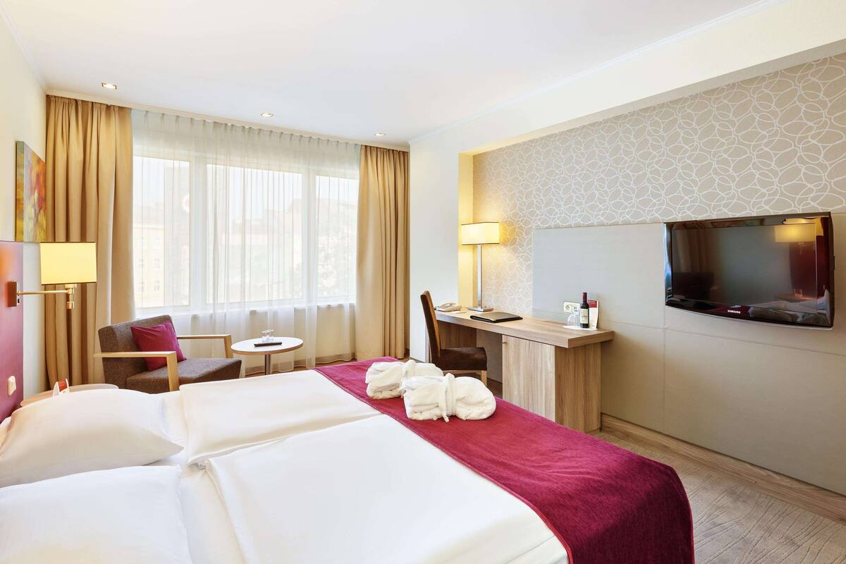 A hotel room with a large bed, a desk with a chair, a TV, and a window with curtains.