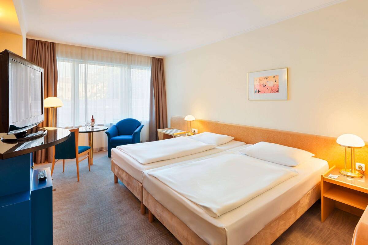 A hotel room with two beds, a desk with a television, a chair, a small table, and a window with curtains.