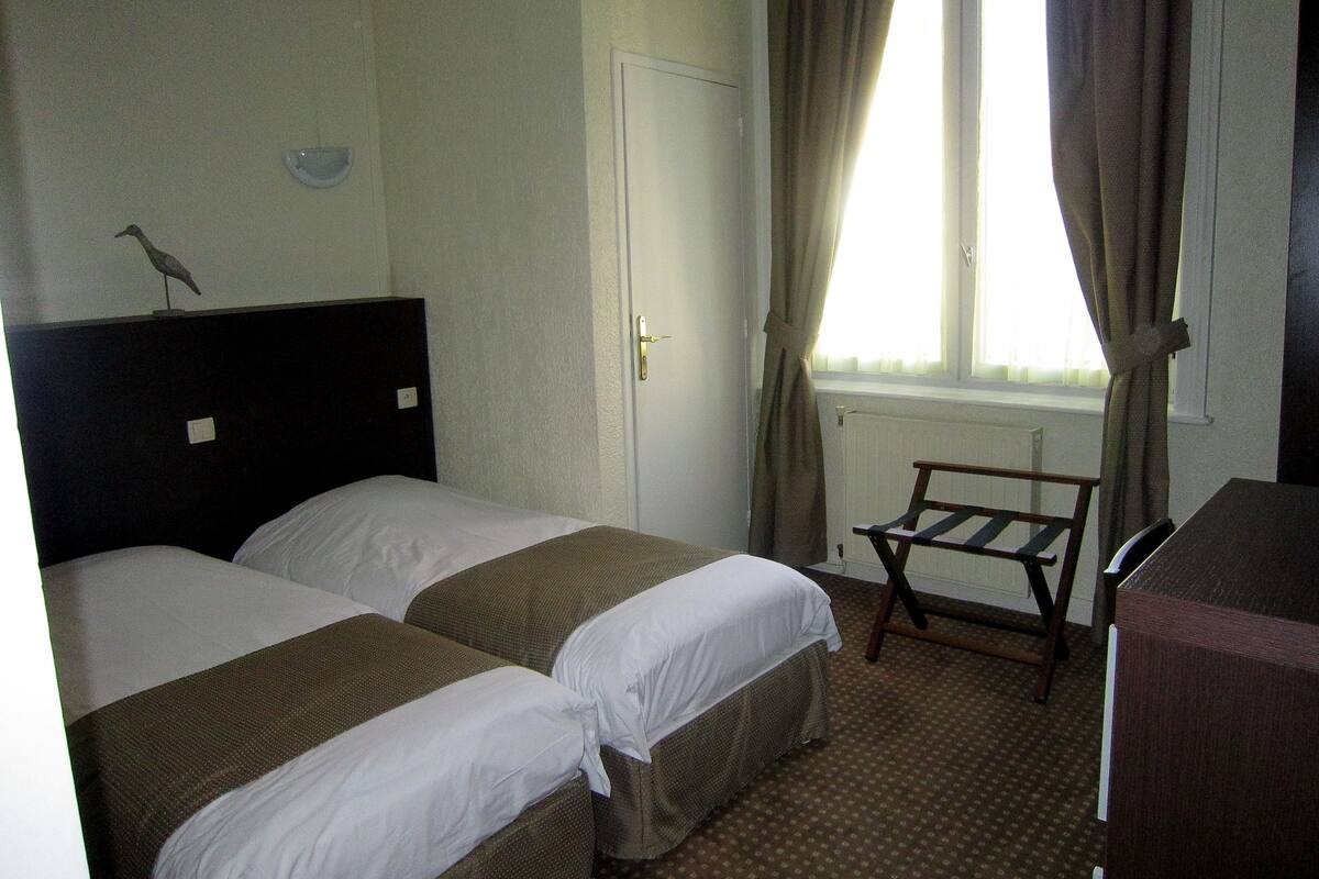 Ein Hotelzimmer mit zwei Einzelbetten, einer Holzbank, einem Heizkörper und einem Fenster mit Vorhängen.