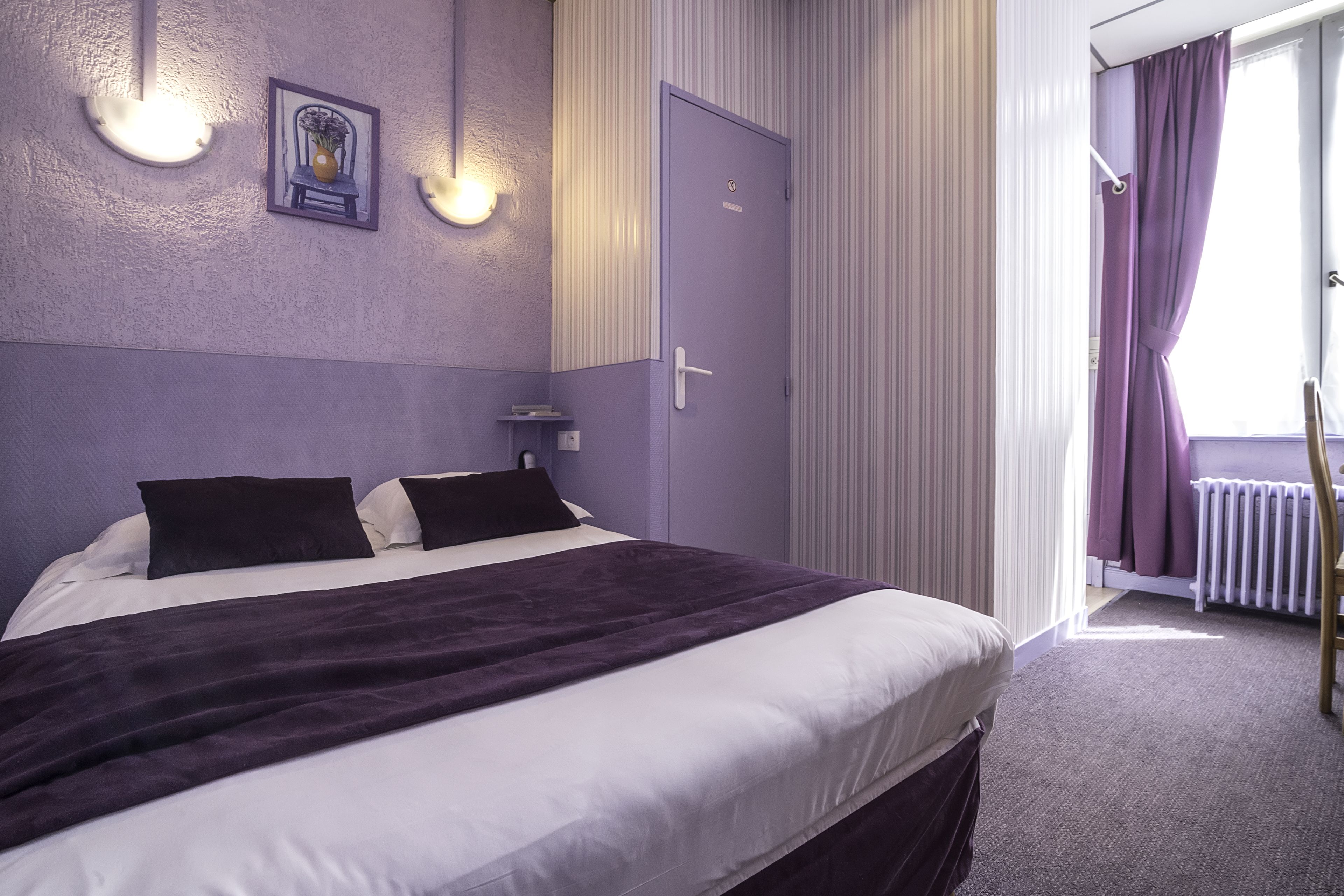 Un dormitorio con una cama, una mesita de noche, una lámpara de pared, una puerta y una ventana con cortinas.