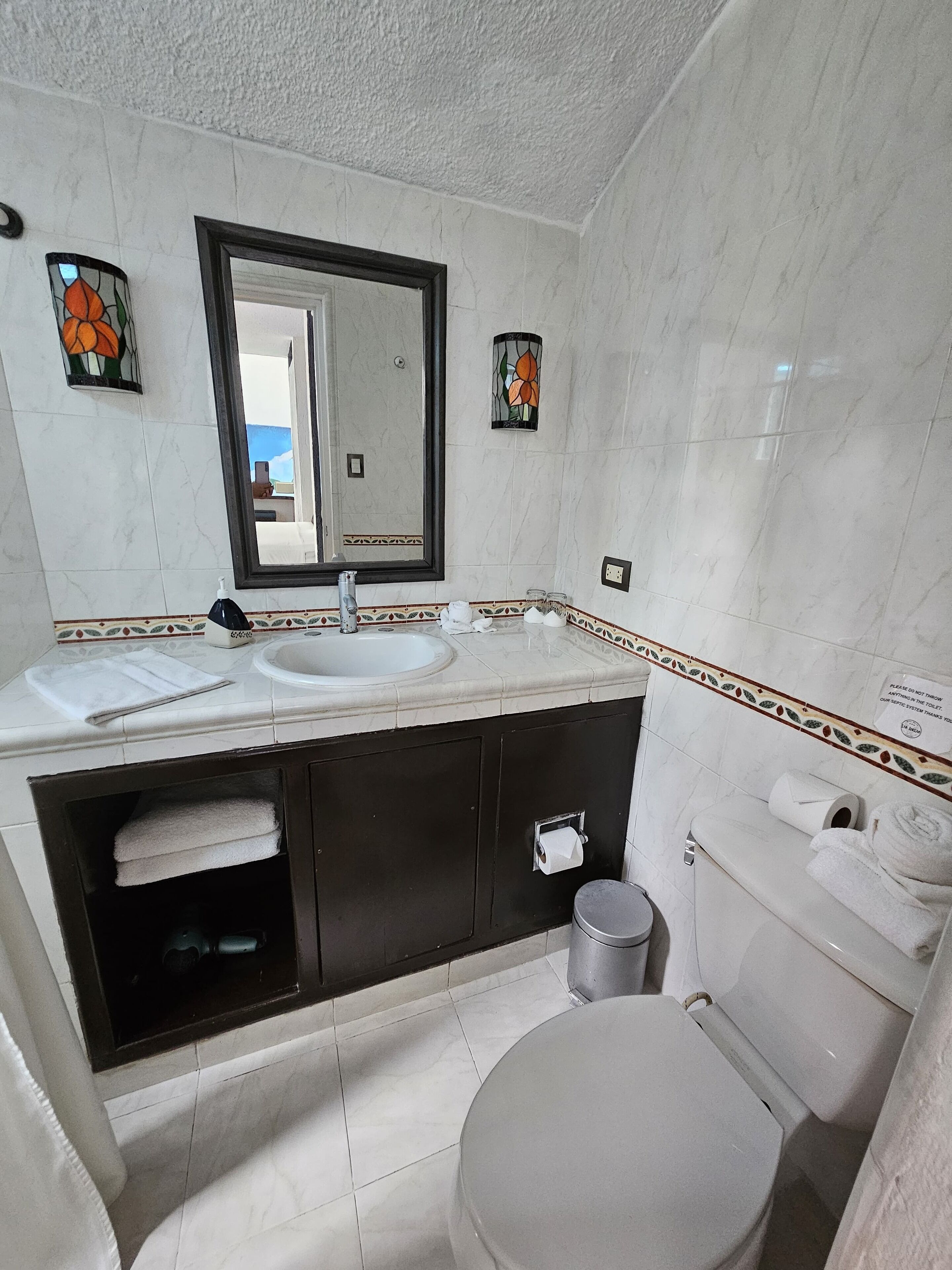 A modern bathroom with a white sink, mirror, and toilet. There are decorative tiles on the walls and a trash can.