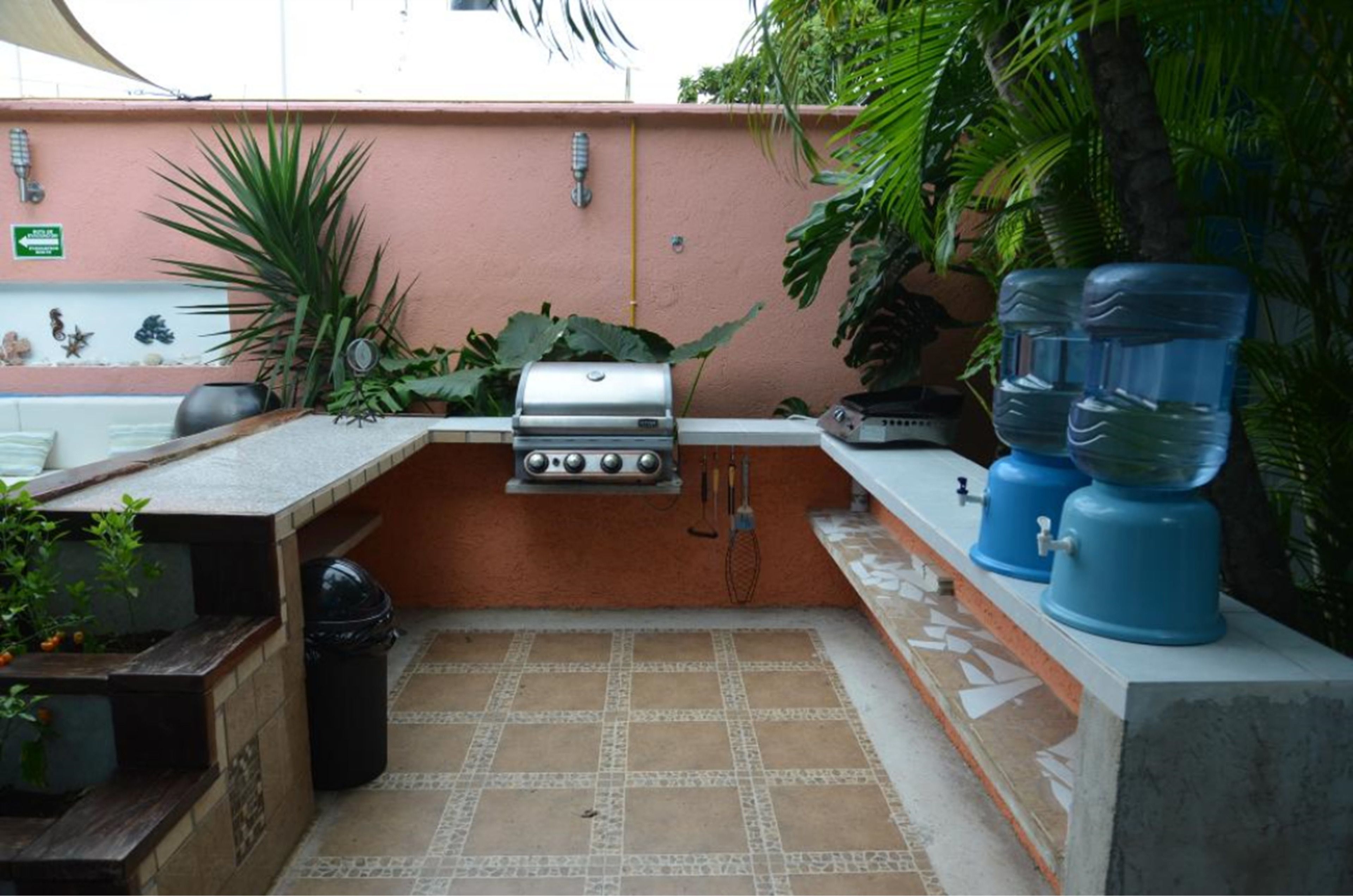 Una cocina exterior compacta con una parrilla a gas, dispensadores de agua y plantas en macetas.