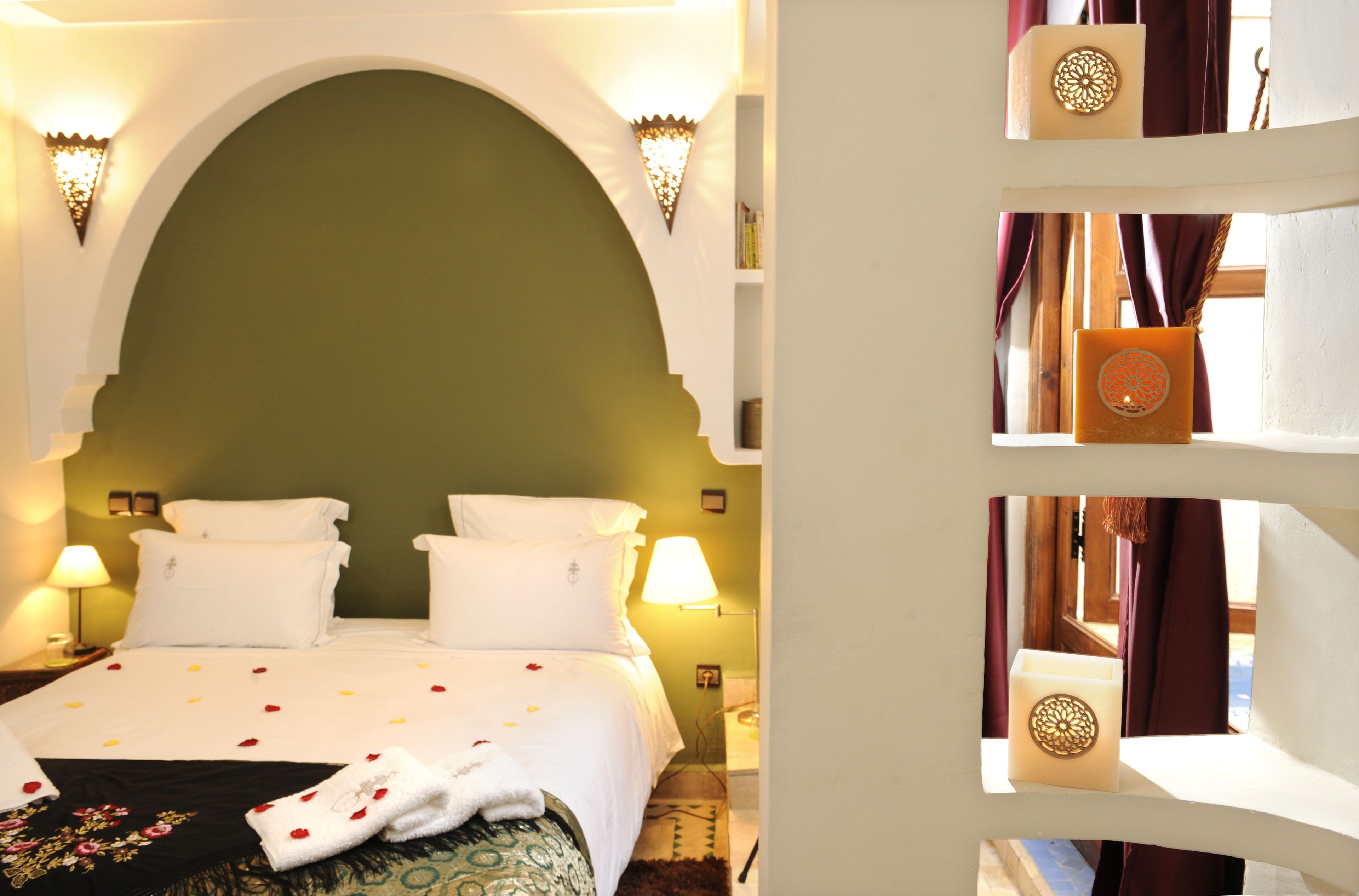 Une chambre d’hôtel avec un grand lit, deux tables de chevet, une petite table d’appoint, une arche décorative sur le mur et une fenêtre avec des rideaux.