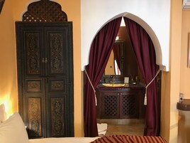 Una habitación con una puerta tradicional, un lavamanos pequeño con espejo y una cama con cubrecolcha a rayas.