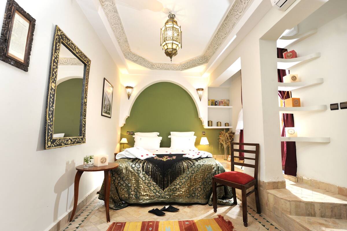 Un dormitorio con un arco verde, una cama con ropa de cama blanca, una silla de madera y una mesita de noche con una lámpara.