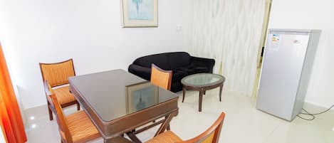 A dining area with a glass-top table, wooden chairs, a black sofa, a small round table, and a refrigerator.