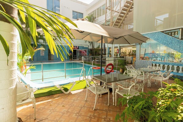 Un espace piscine intérieur doté d’un toit en verre, de chaises blanches et de tables placées sous un grand parasol.
