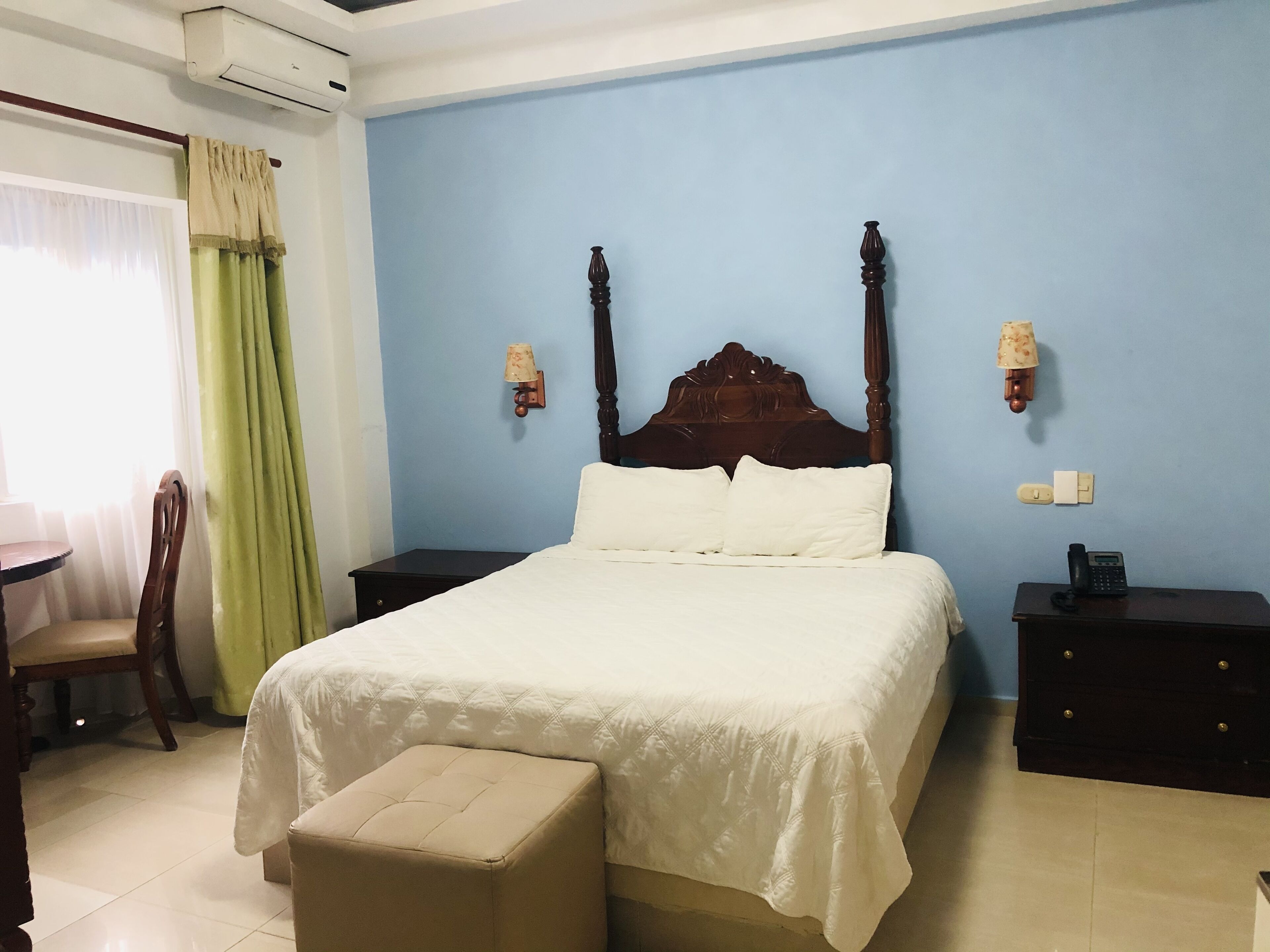 Un dormitorio con una cama con dosel de madera, una mesita de noche con lámpara, una silla y un pequeño reposapiés.