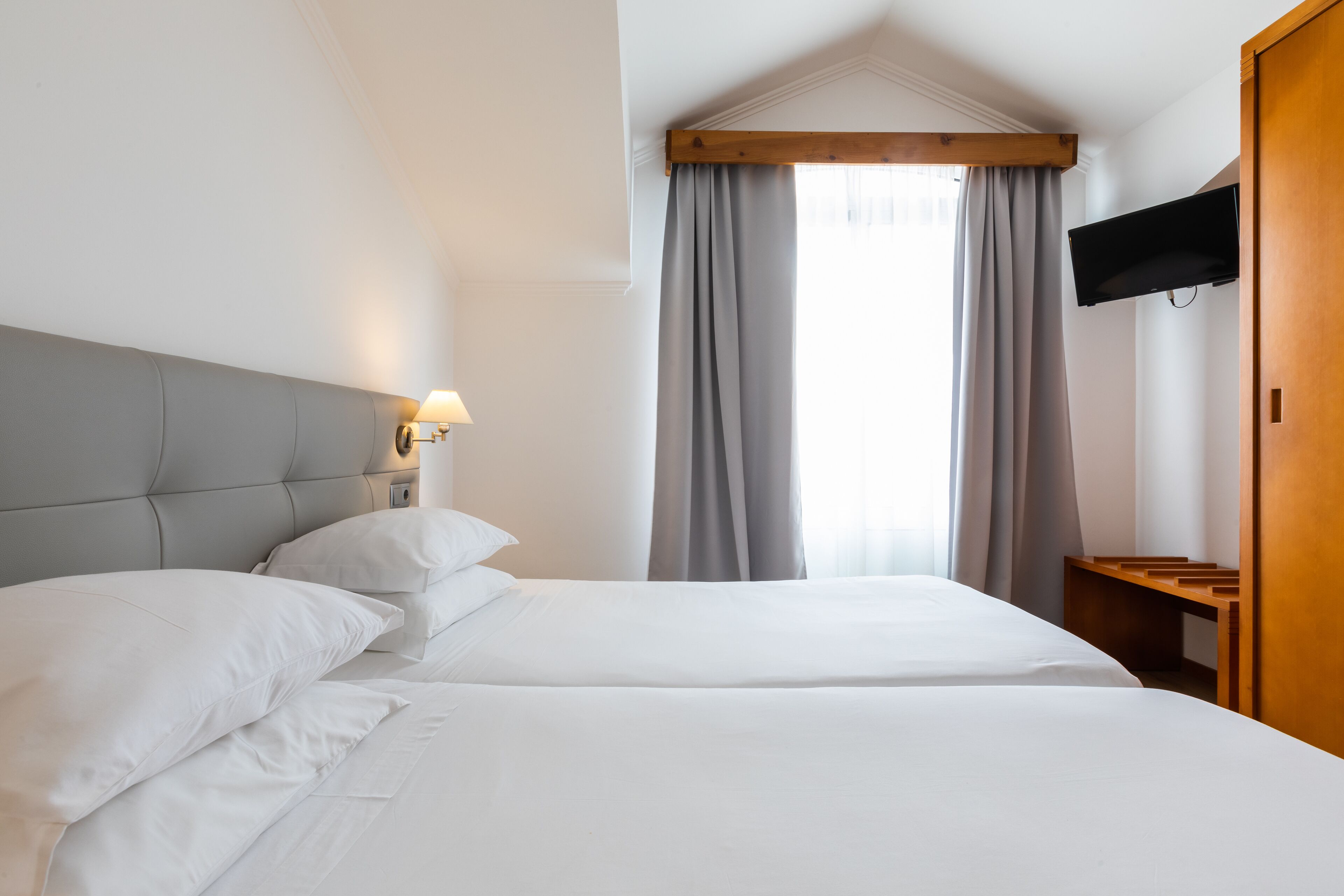 Une chambre d’hôtel avec deux lits, un téléviseur fixé au mur et un banc en bois.