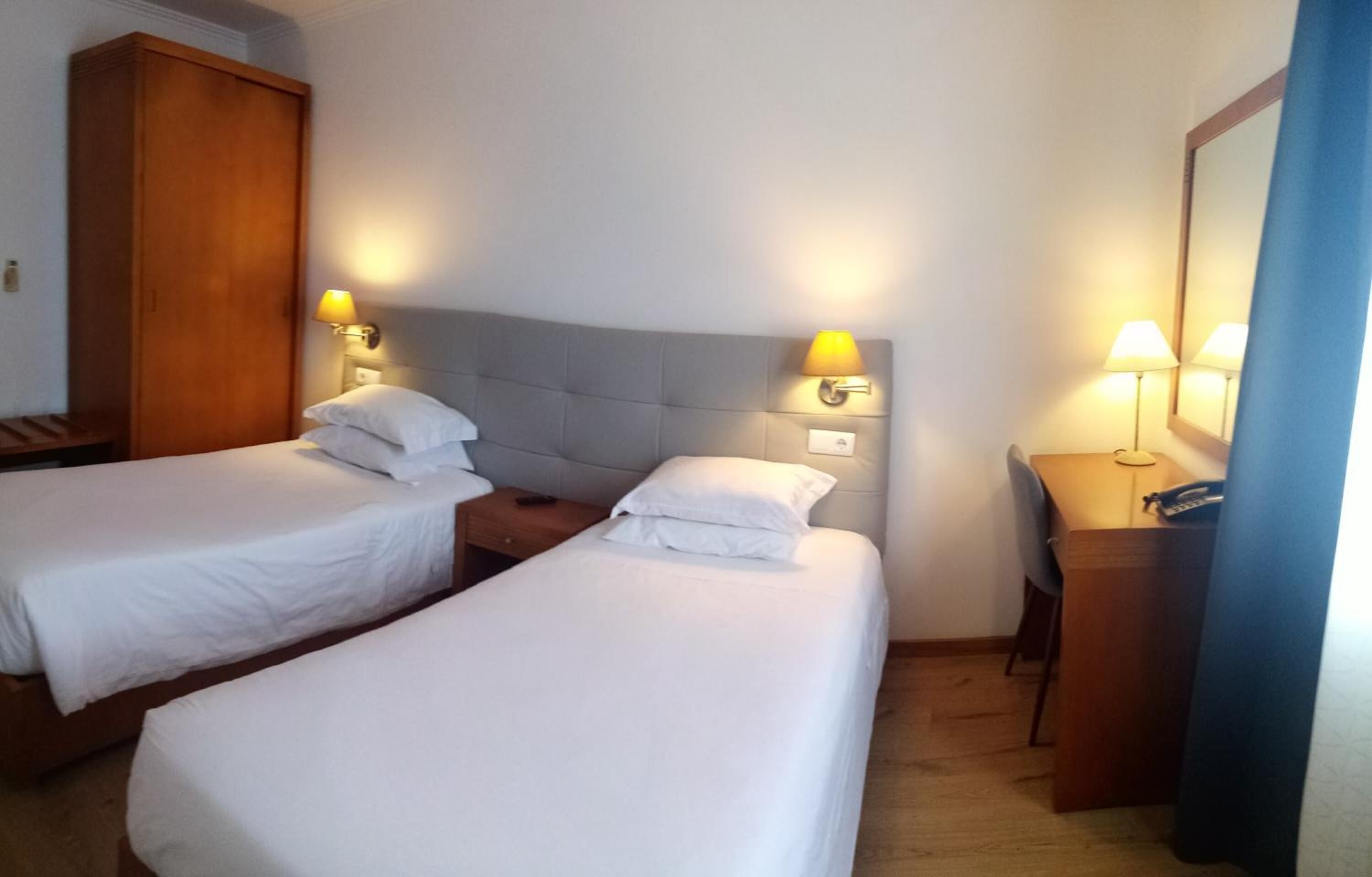 A hotel room with two single beds, a wooden wardrobe, a desk with a lamp, and a mirror.