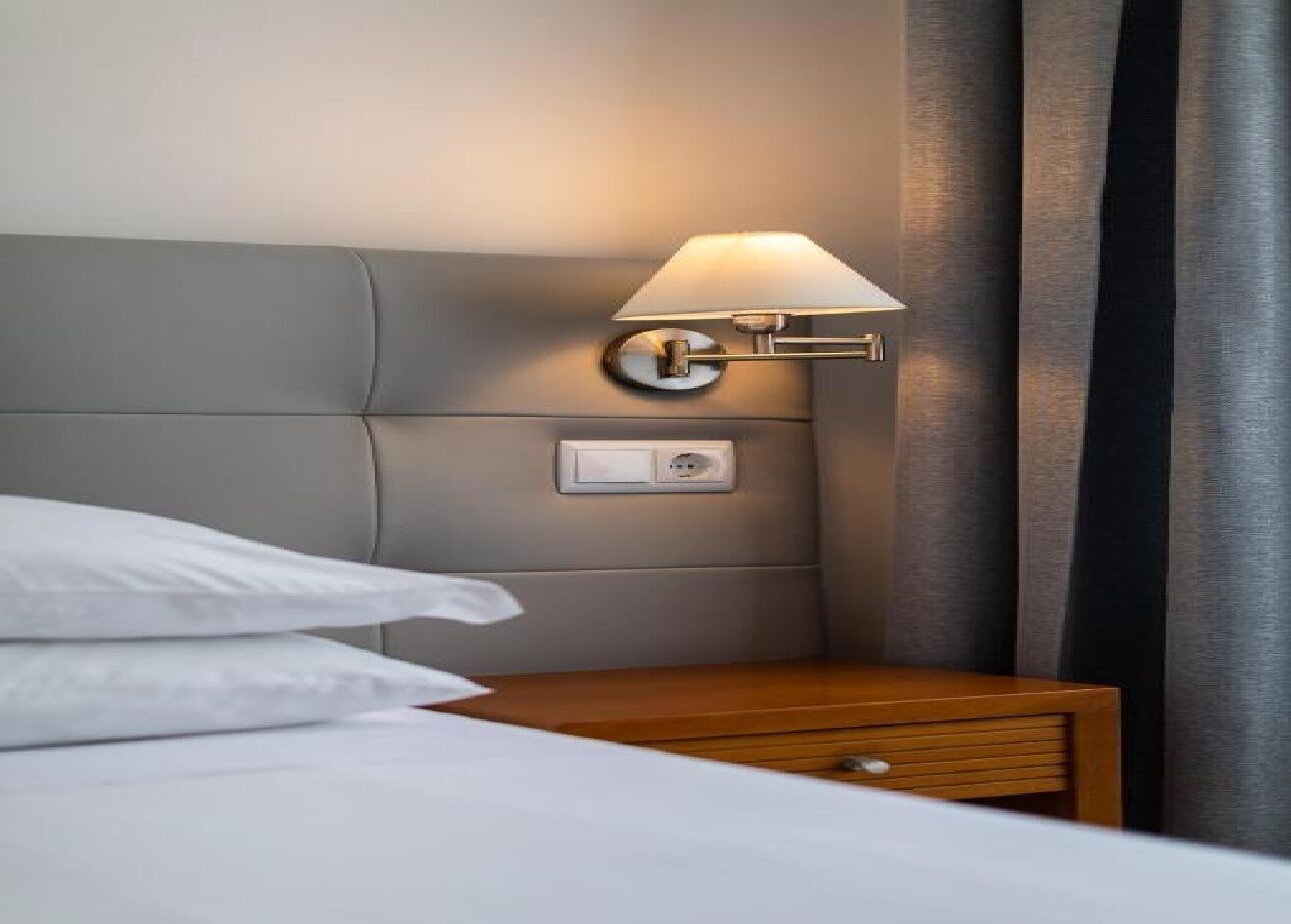 Un lit bien fait, avec du linge de lit blanc, une table de chevet en bois avec une lampe et une prise électrique sur le mur.