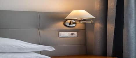 A neatly made bed with white linens, a wooden nightstand with a lamp, and a power outlet on the wall.