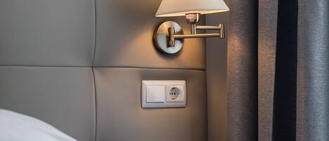 A neatly made bed with white linens, a wooden nightstand with a lamp, and a wall socket.