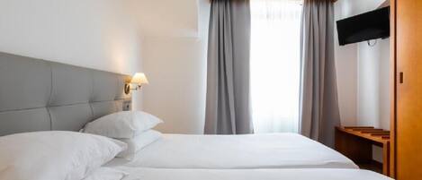 A hotel room with two beds, a TV mounted on the wall, a wooden headboard, and a window with curtains.