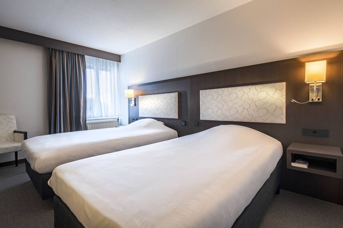 A hotel room with two beds, a chair, a lamp, and a wall-mounted shelf.