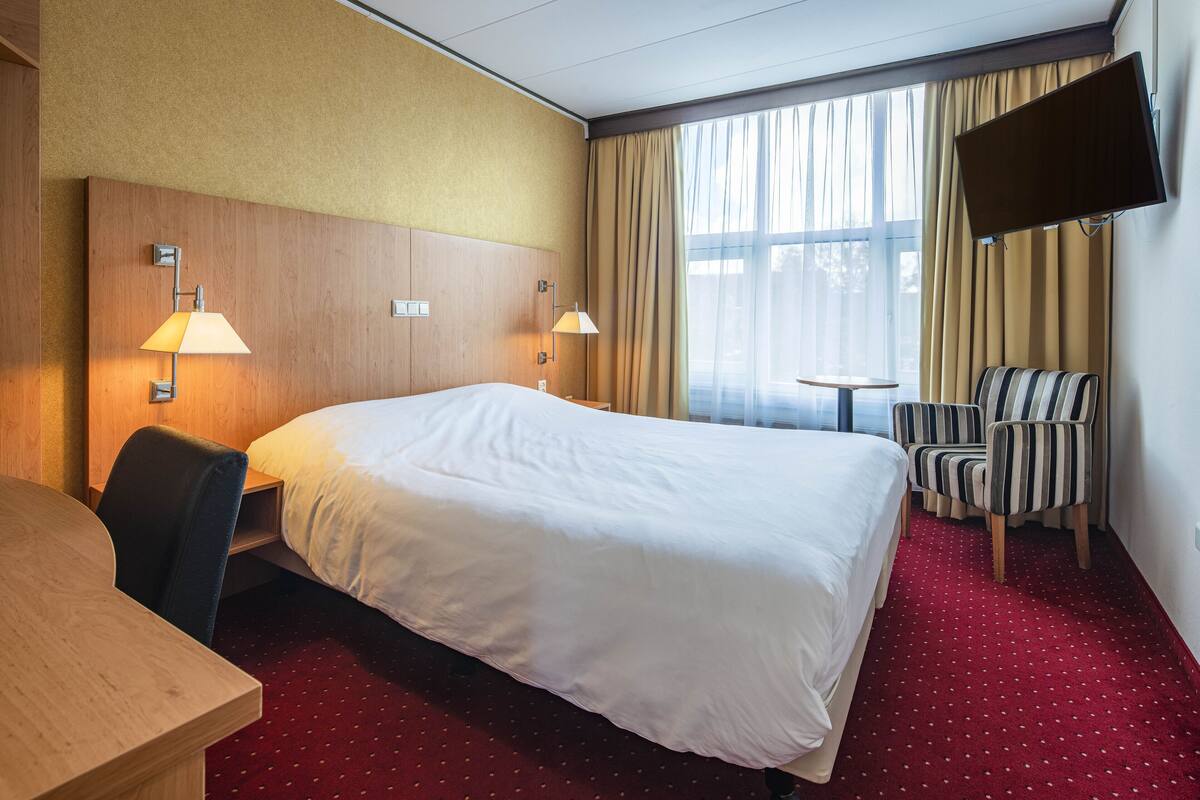 A hotel room with a large bed, two bedside lamps, a TV mounted on the wall, a desk, and a striped chair.