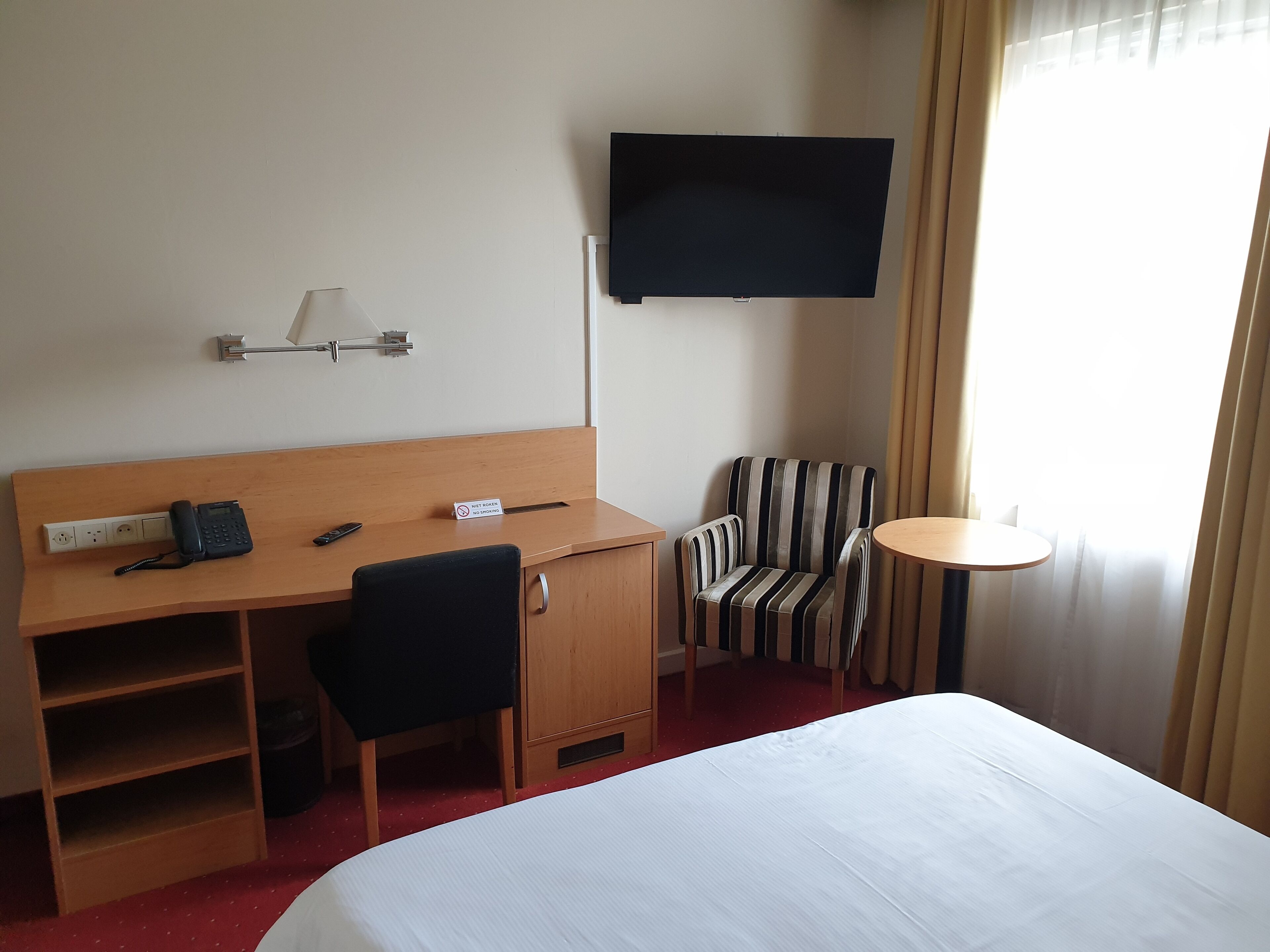 A hotel room with a bed, a desk with a telephone, a chair, a television mounted on the wall, and a small table.