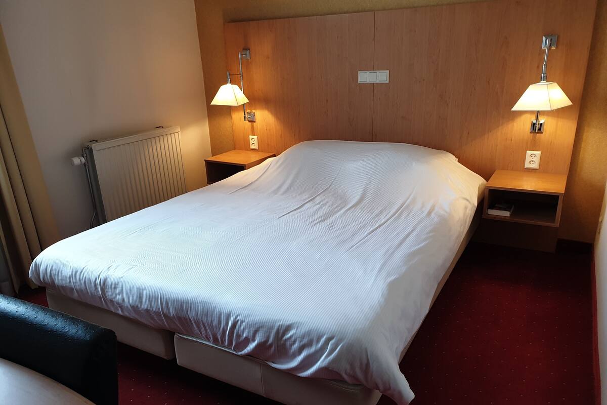 A neatly made bed with a white comforter and pillows, flanked by wooden nightstands with lamps, in a room with a wooden headboard and a radiator.