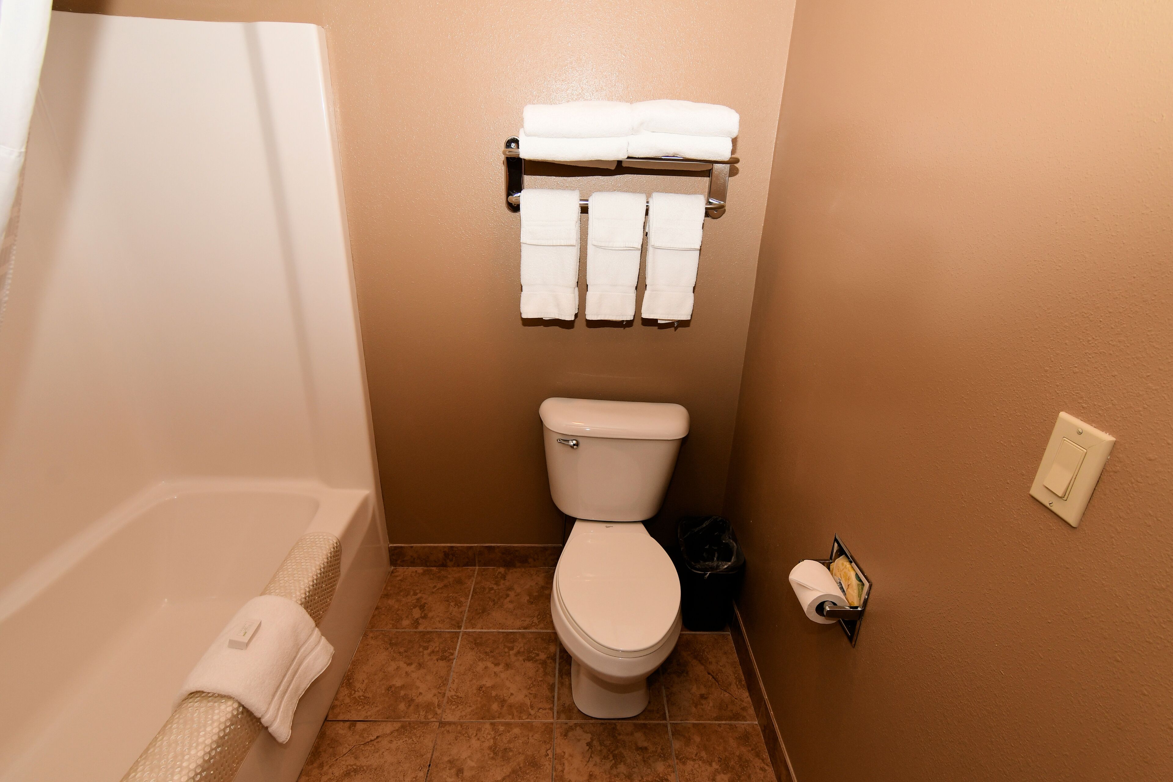 A bathroom with a toilet, a towel rack with towels, a toilet paper holder, and a trash bin.