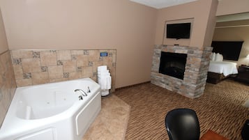 A hotel room with a large bathtub, a stone fireplace, a flat-screen TV mounted above the fireplace, and a dining area with a table and chairs.