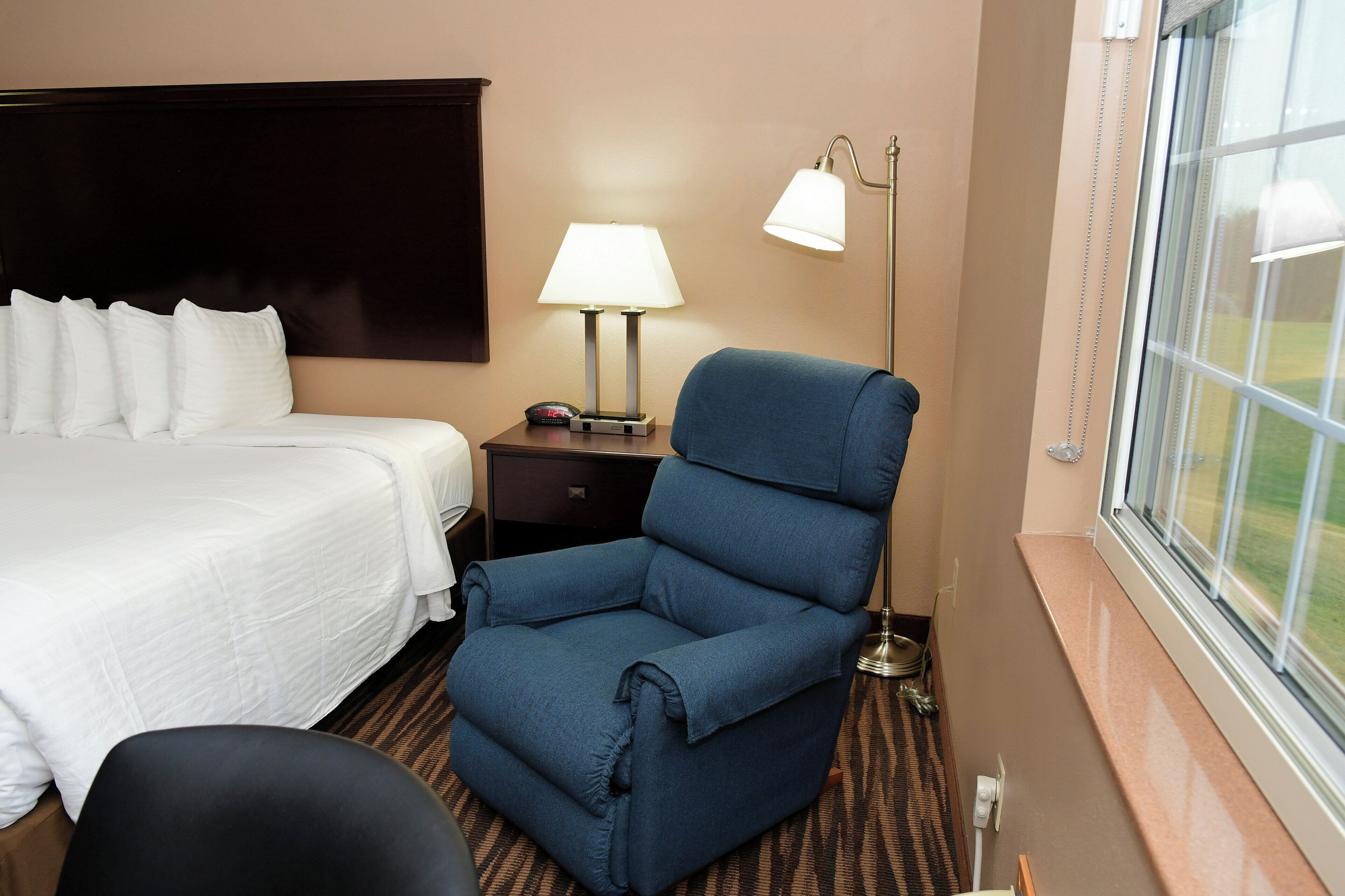 A hotel room with a bed, a blue armchair, a nightstand with a lamp, and a window with a view.