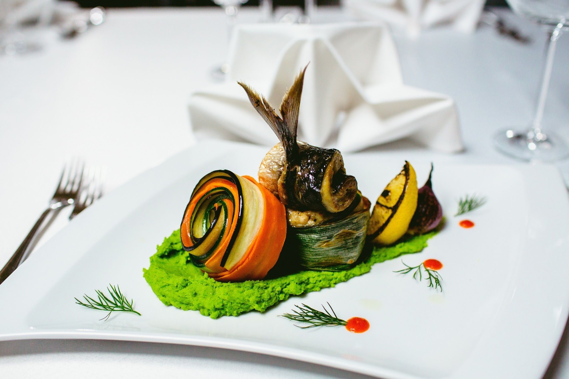 Un plato blanco con puré verde como base, un vegetal enrollado y un pescado decorado.