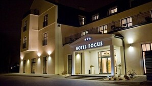 Un hotel de dos pisos llamado "HOTEL FOCUS", con fachada moderna, ventanas iluminadas y un balcón en el piso superior.