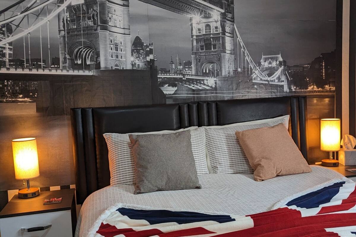 A hotel room with a large bed, a headboard, two bedside tables with lamps, a nightstand, and a wall mural of a bridge.