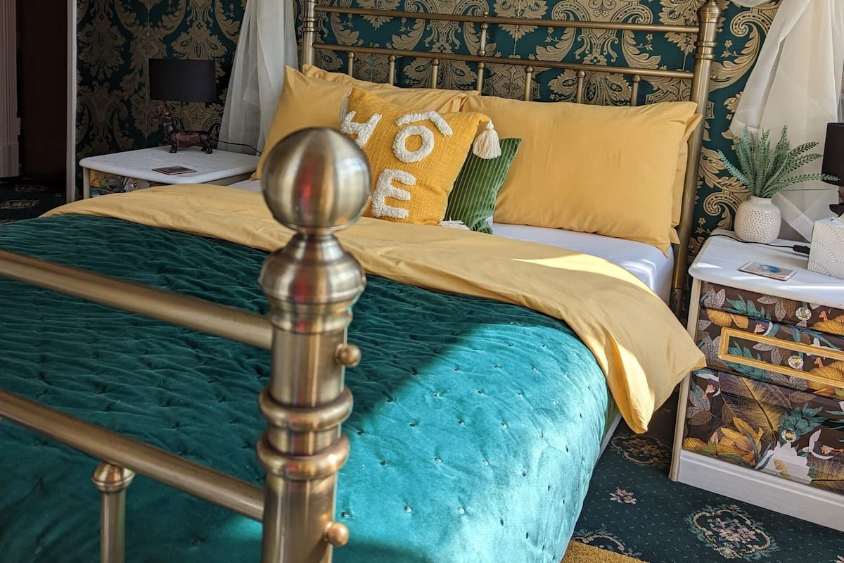 A bed with a teal bedspread, a gold headboard, and yellow pillows. There is a painting above the bed and a nightstand with a lamp and a plant.