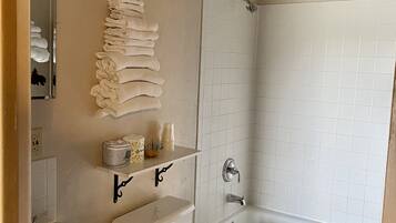 A bathroom with a toilet, sink, and bathtub. There are towels stacked on a shelf, and a shower curtain rod is visible.