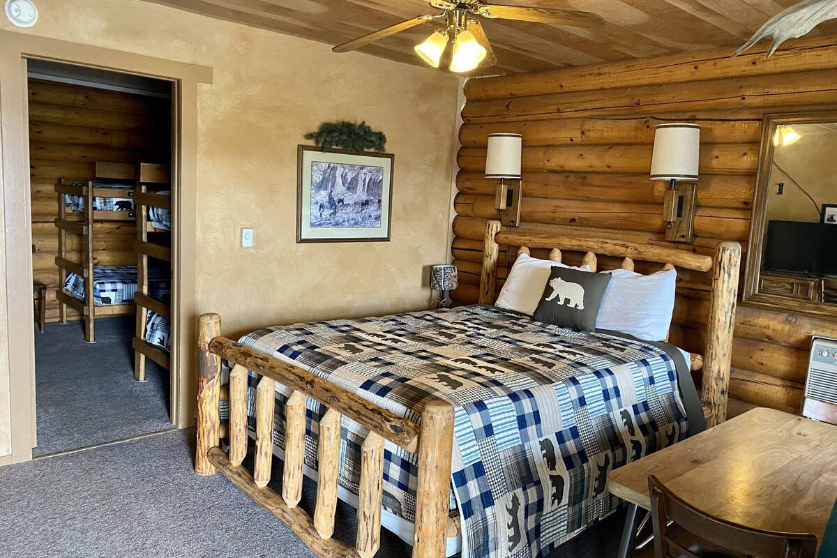 Un dormitorio tipo cabaña de troncos con una cama de madera, un ventilador de techo, un cuadro enmarcado en la pared y una mesa de madera con sillas.