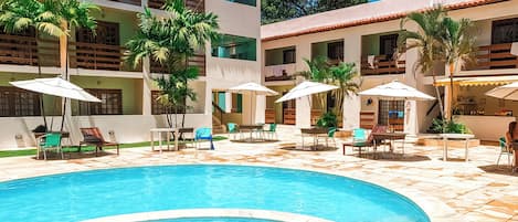 Una piscina con bordes azulejados, rodeada de zonas de estar al aire libre con sombrillas, y un edificio con balcones y ventanas.