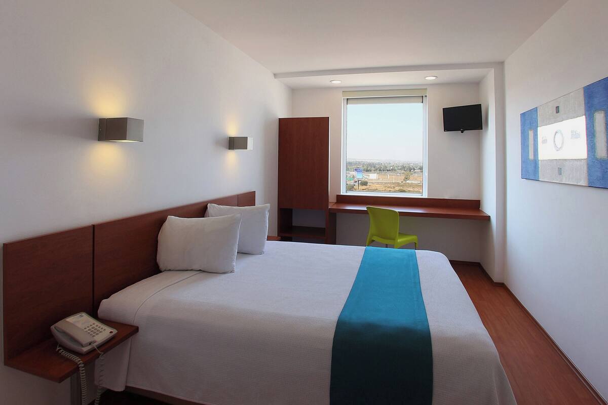 Habitación de hotel con cama, teléfono en la mesita de noche, escritorio, silla, televisión y ventana con vistas.