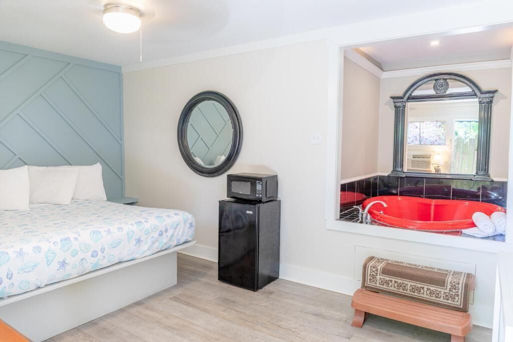 A bedroom with a bed, a small black refrigerator, a round mirror, and a red bathtub.