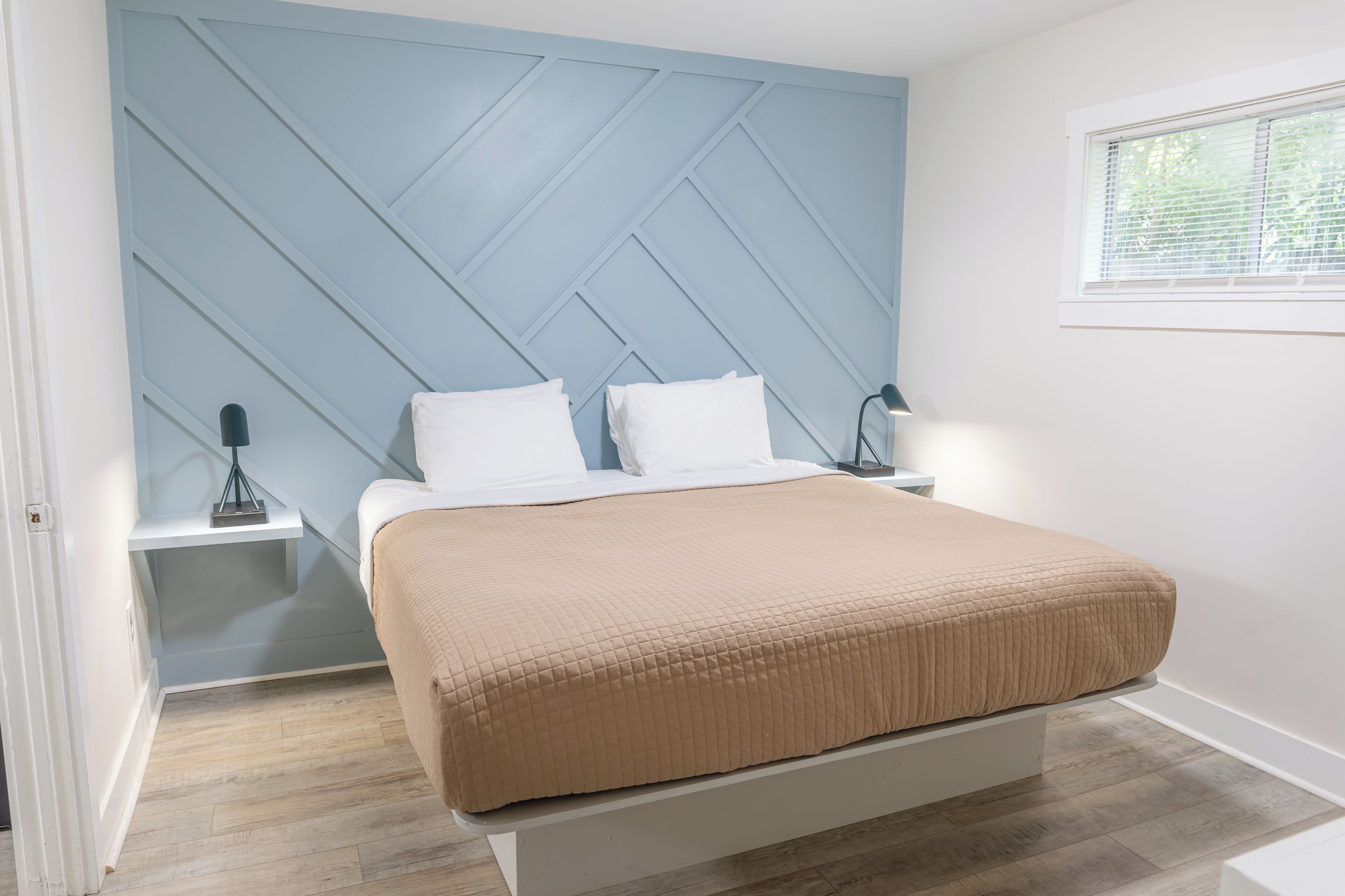 A bedroom with a bed, white nightstands, a wall-mounted lamp, and a window with blinds.