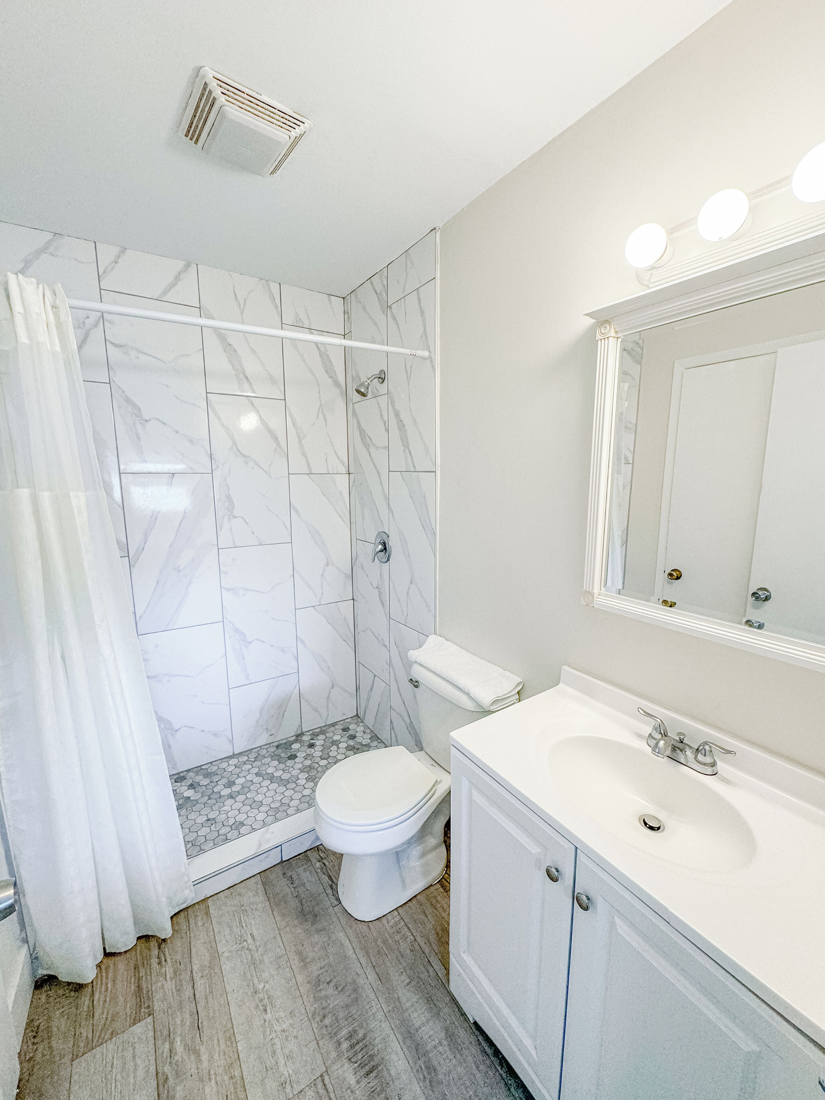 A bathroom with a shower, toilet, and sink, featuring marble tiles and a wooden floor.
