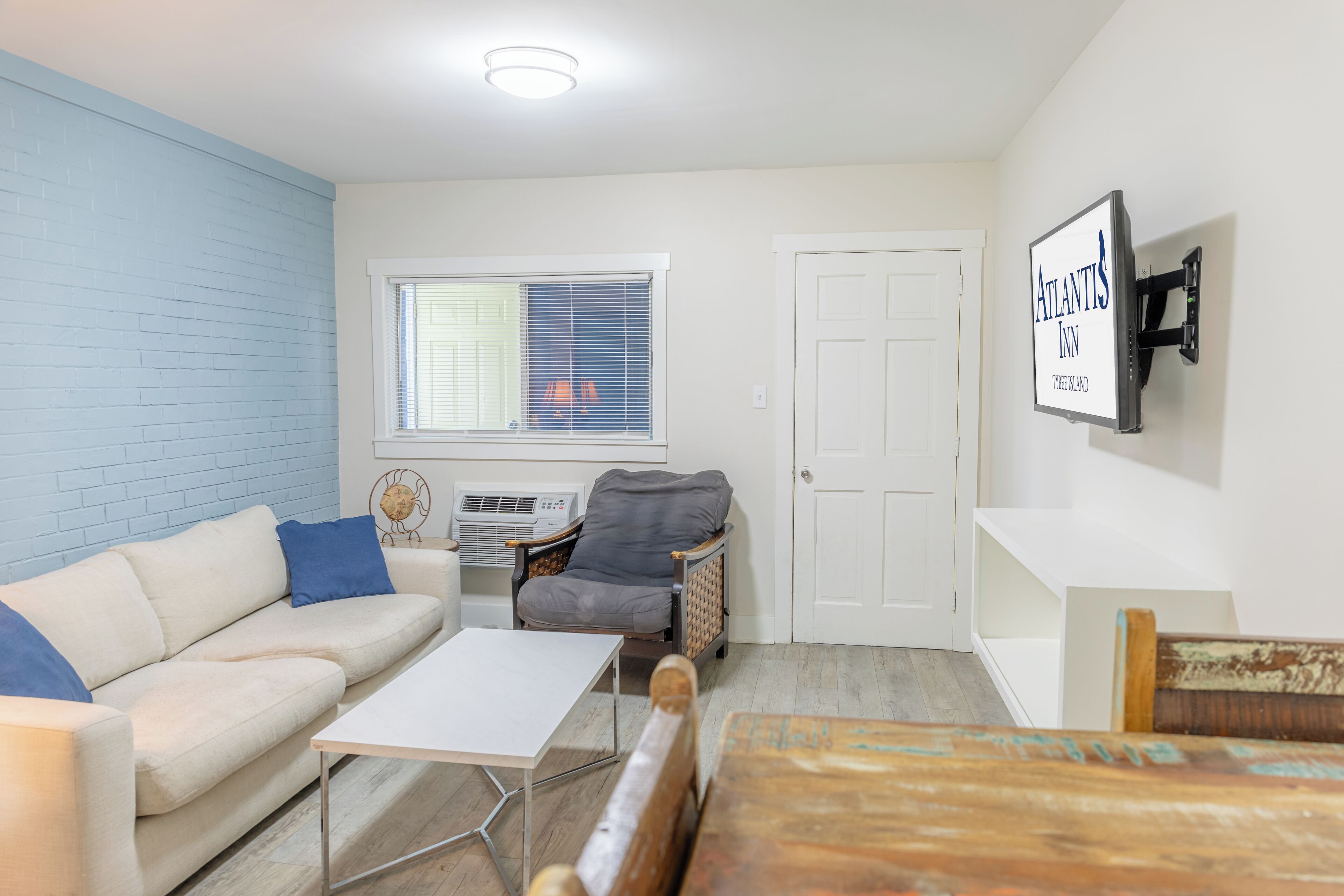 A living room with a white sofa, a small table, a wicker chair, a TV mounted on the wall, and a window with blinds.