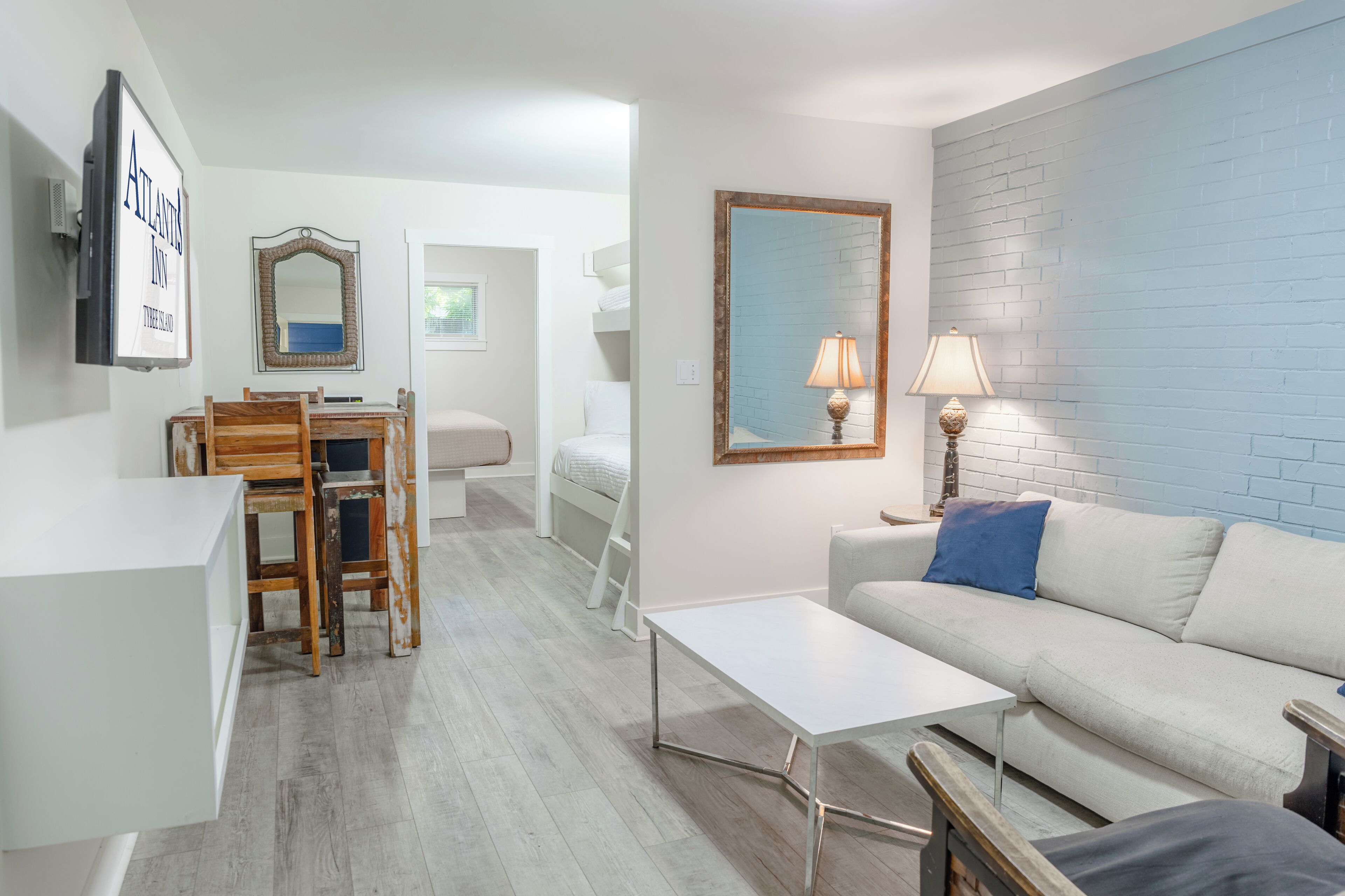 A modern living room with a white couch, a small white table, a wooden chair, a dining table with chairs, a mirror, and a lamp.
