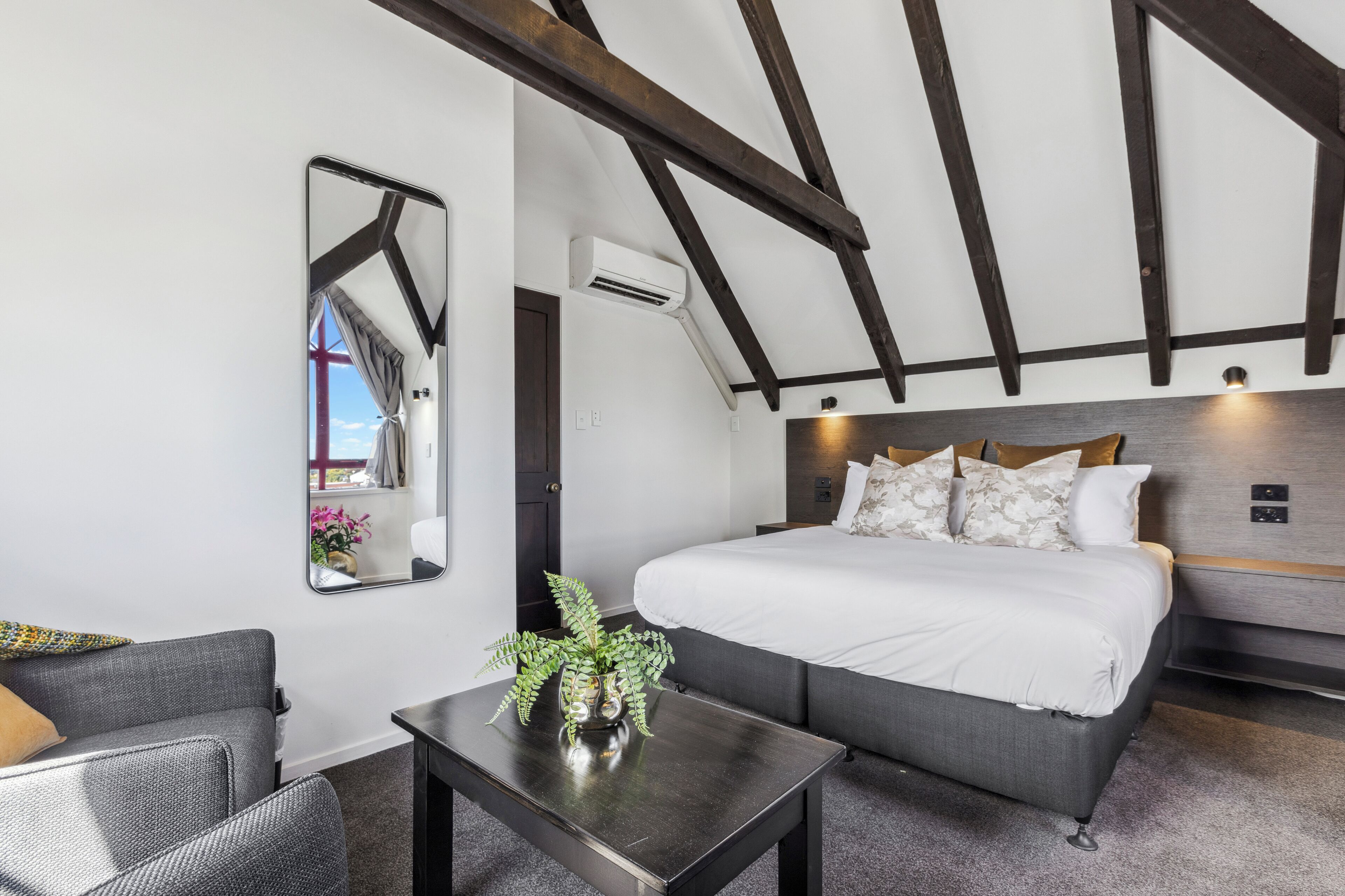 A modern bedroom with a large bed, a small table with a plant, a mirror, and a view of the outdoors.