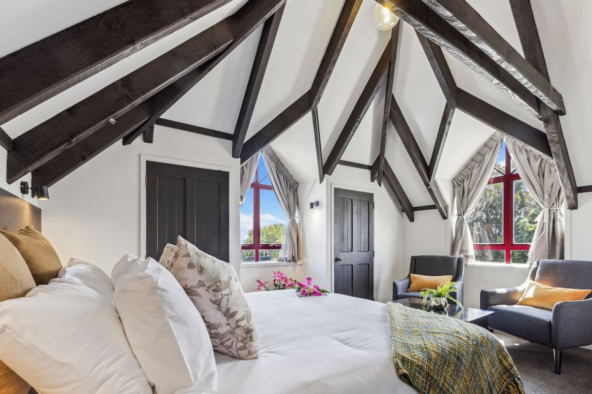 Un dormitorio con una cama grande, dos sillones, una mesita y una ventana con cortinas.