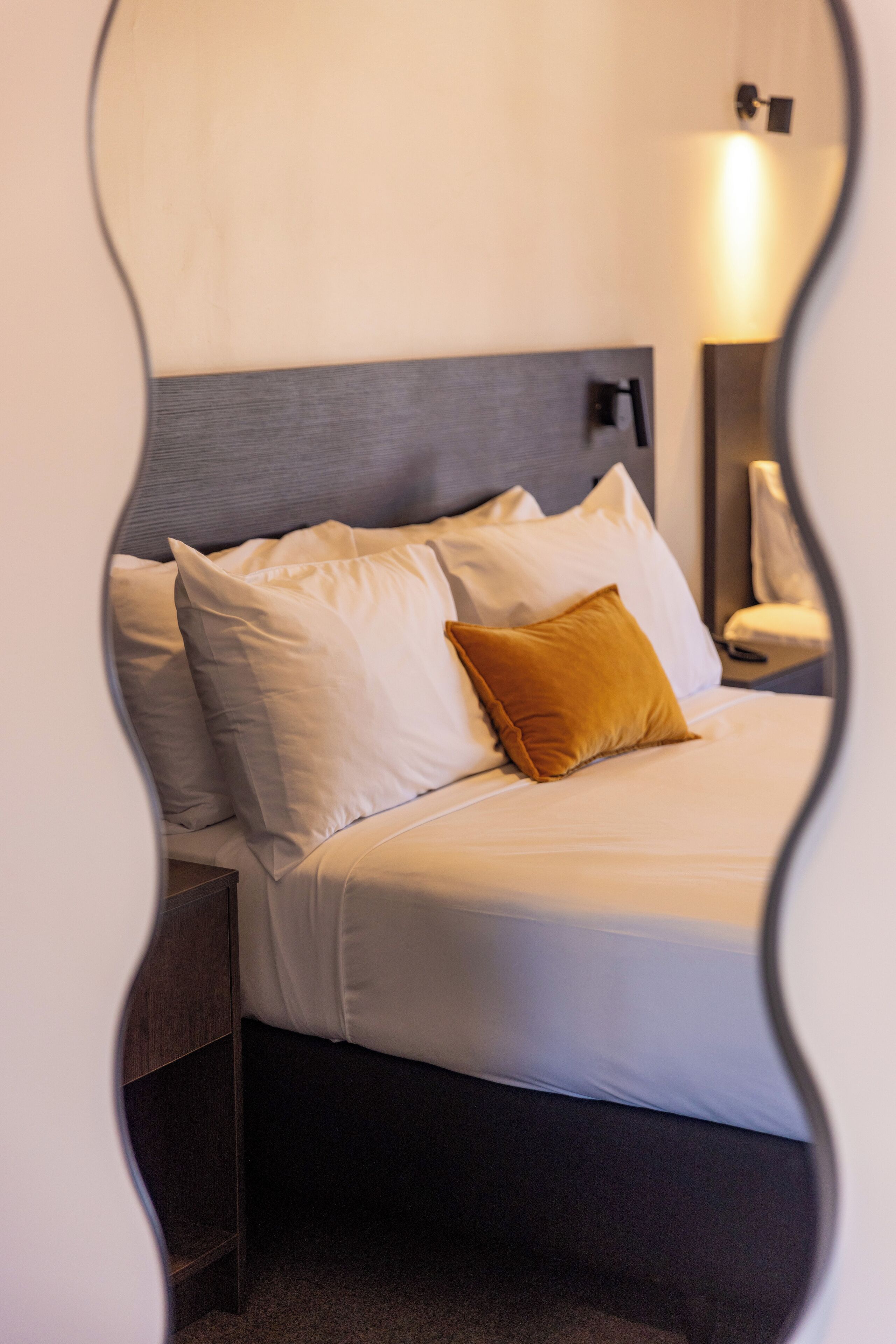 A neatly made bed with white and mustard pillows, a headboard, and a bedside table with a lamp.