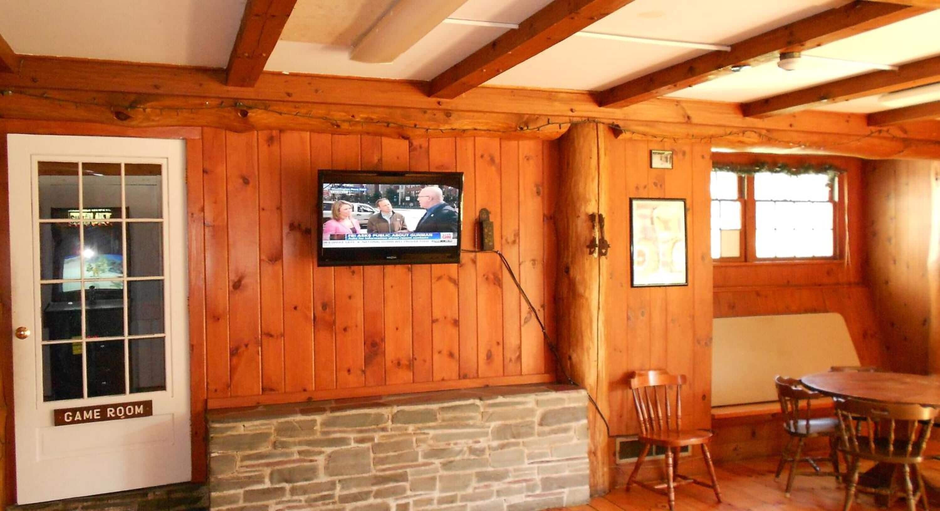 A wooden-paneled room with a television mounted on the wall, a stone fireplace, a door labeled "GAME ROOM," and a dining area with wooden chairs and a table.