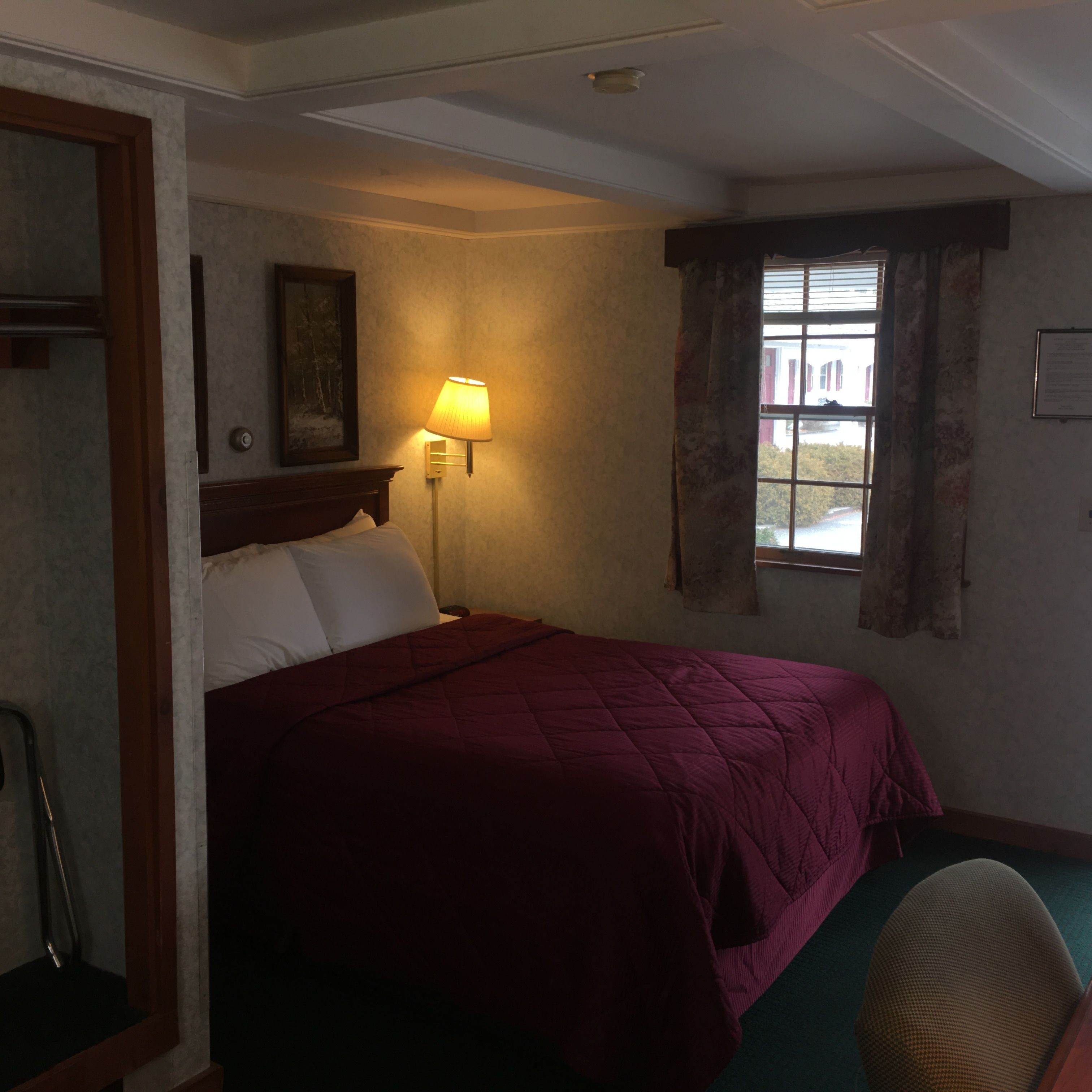 A hotel room with a bed, a nightstand, a lamp, a window with curtains, and a framed picture on the wall.