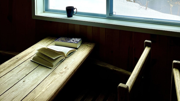A wooden cabin interior with a window overlooking a snowy landscape, a wooden table with books and a mug, and a chair. - Stonybrook Motel & Lodge (Franconia)