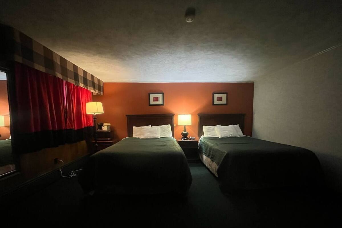A hotel room with two single beds, a nightstand with a lamp, and a window with red curtains.