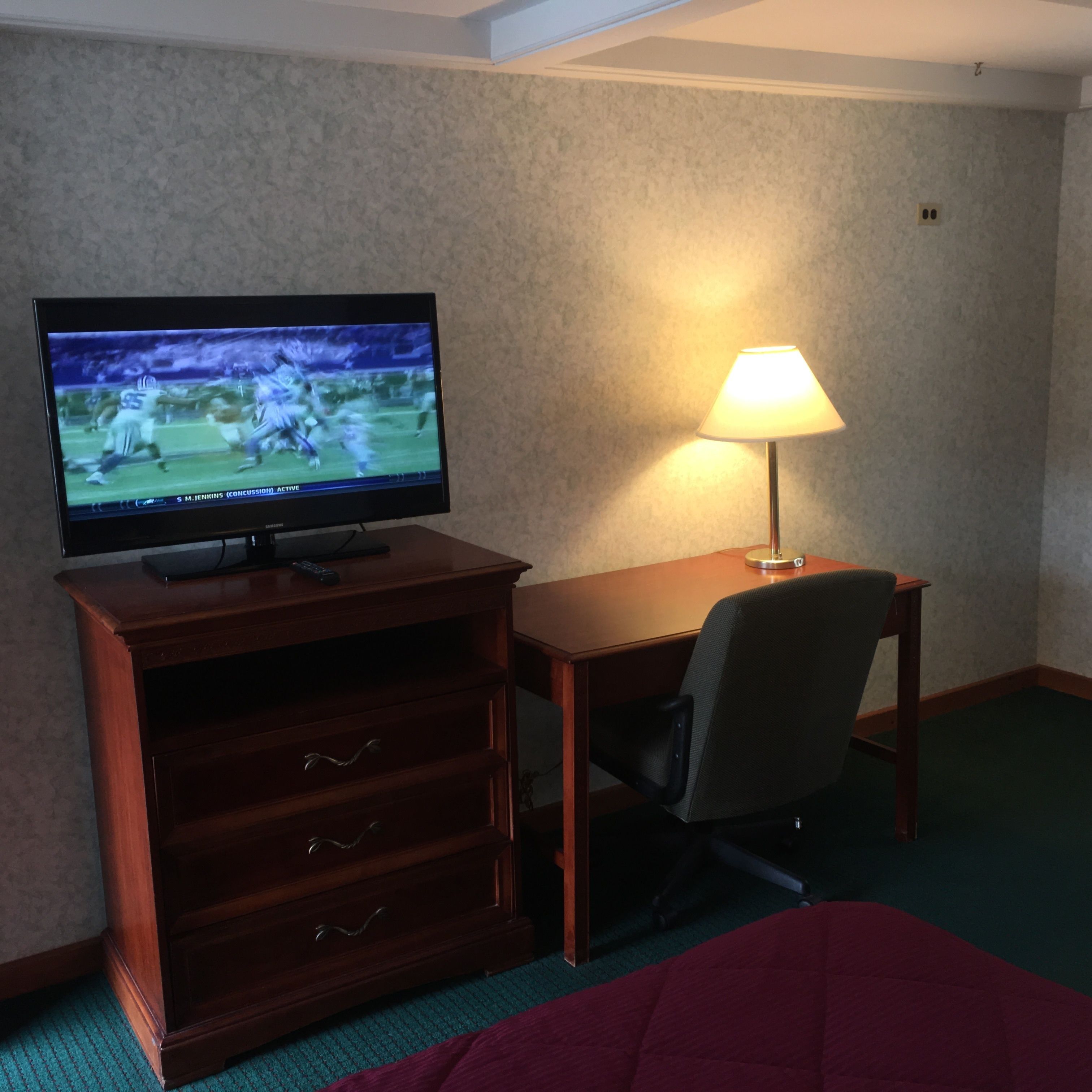 A hotel room with a television on a wooden cabinet, a desk with a lamp, a chair, and a bed with a red blanket.