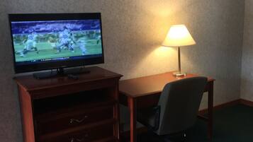 A hotel room with a television on a wooden cabinet, a desk with a lamp, a chair, and a bed with a red blanket.