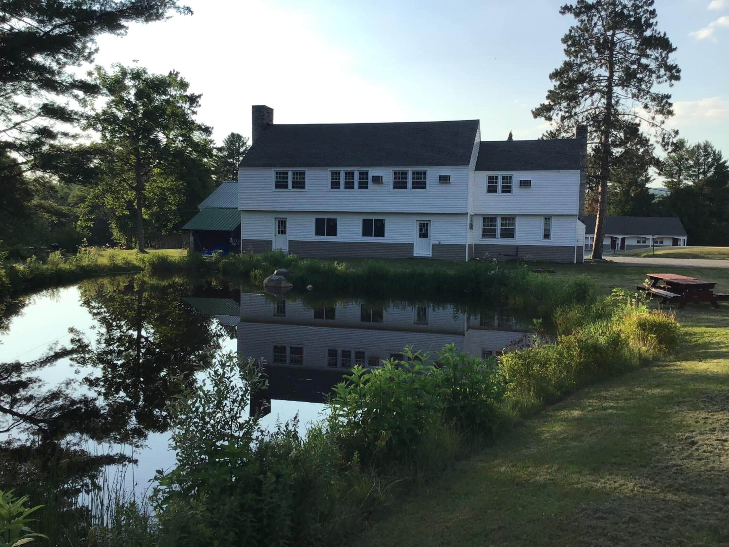 En vit byggnad i två våningar med en symmetrisk fasad, en liten damm framför och ett picknickbord till höger.