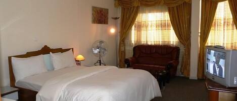 A hotel room with a large bed, a leather armchair, a television, and a fan.