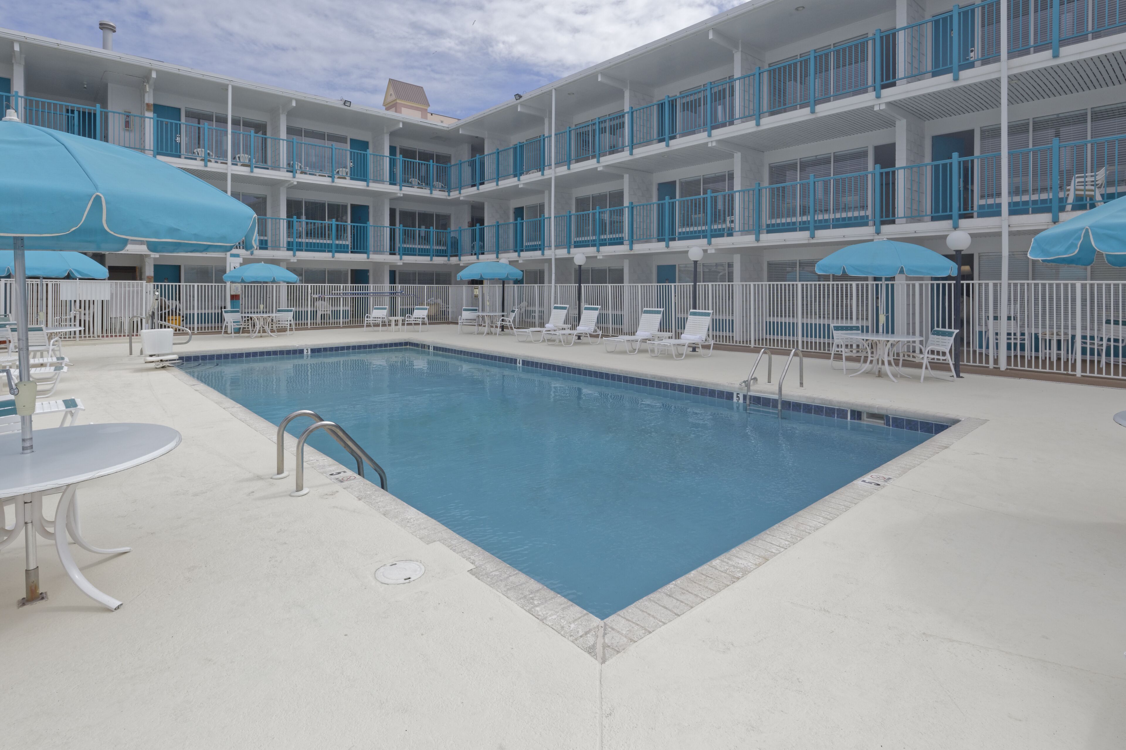 A swimming pool with lounge chairs and umbrellas, surrounded by a white fence and a multi-story building with balconies.